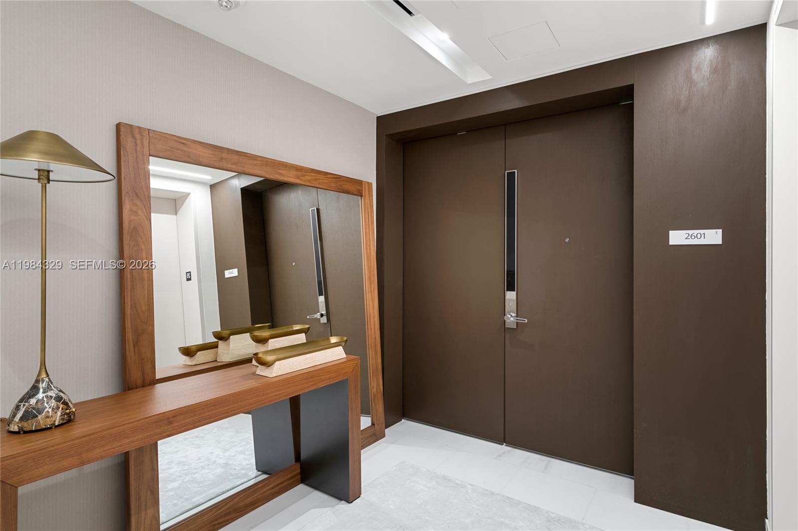 A modern entryway features a wooden console table, a large mirror, and double brown doors — 300 Biscayne Boulevard Way # 2601E, Miami FL 33131, MIAMI, FL