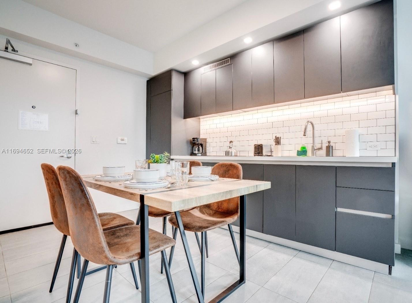 A modern kitchen features dark gray cabinetry, white subway tile backsplash, and a wooden dining table — 227 NE 2nd St # 1705, Miami FL 33132, MIAMI, FL