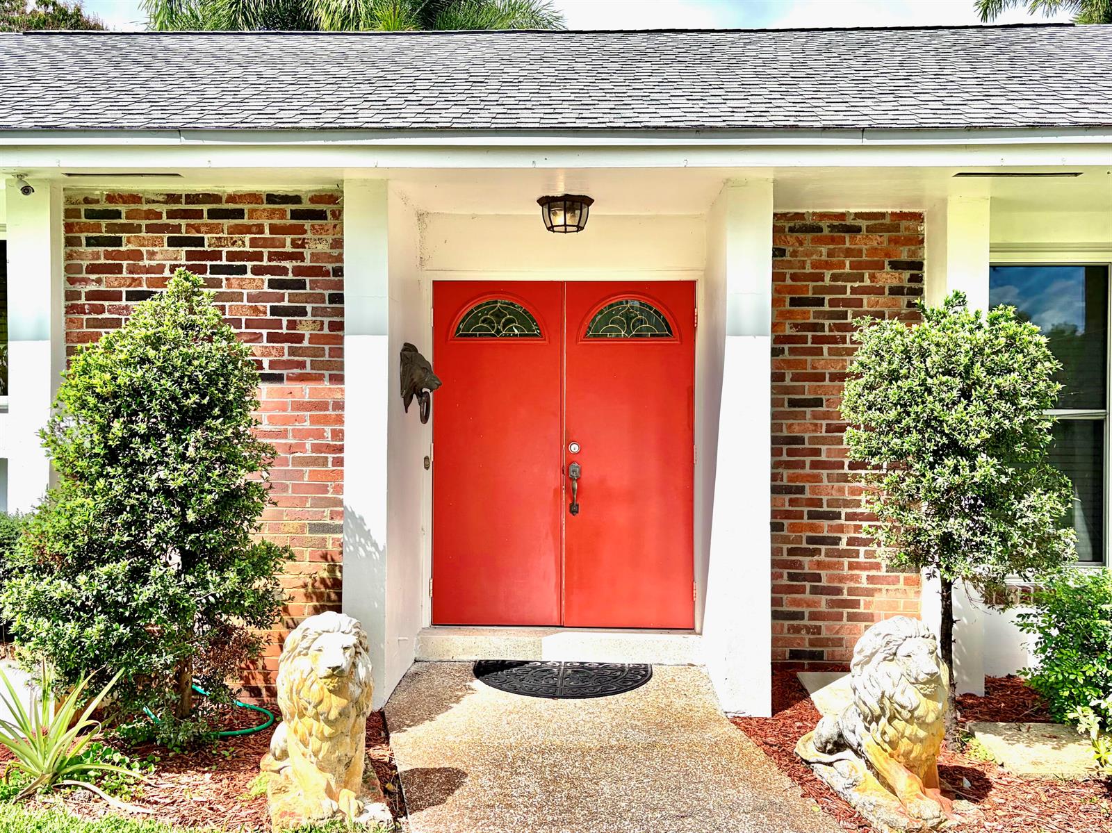 A red double front door is flanked by two lion statues and brick exterior walls — 8 Bell Lane, Palm Springs FL 33406, PALM SPRINGS, FL