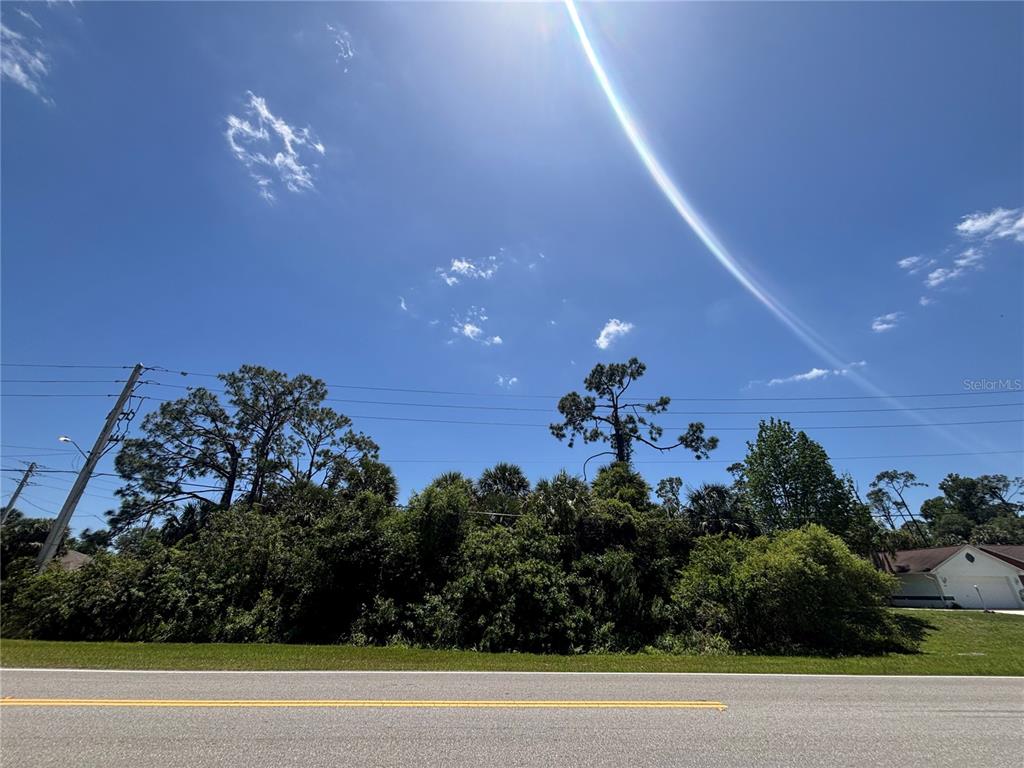 A paved road runs in front of a densely wooded lot under a sunny sky — Lot #11 N CHAMBERLAIN BOULEVARD, NORTH PORT, FL