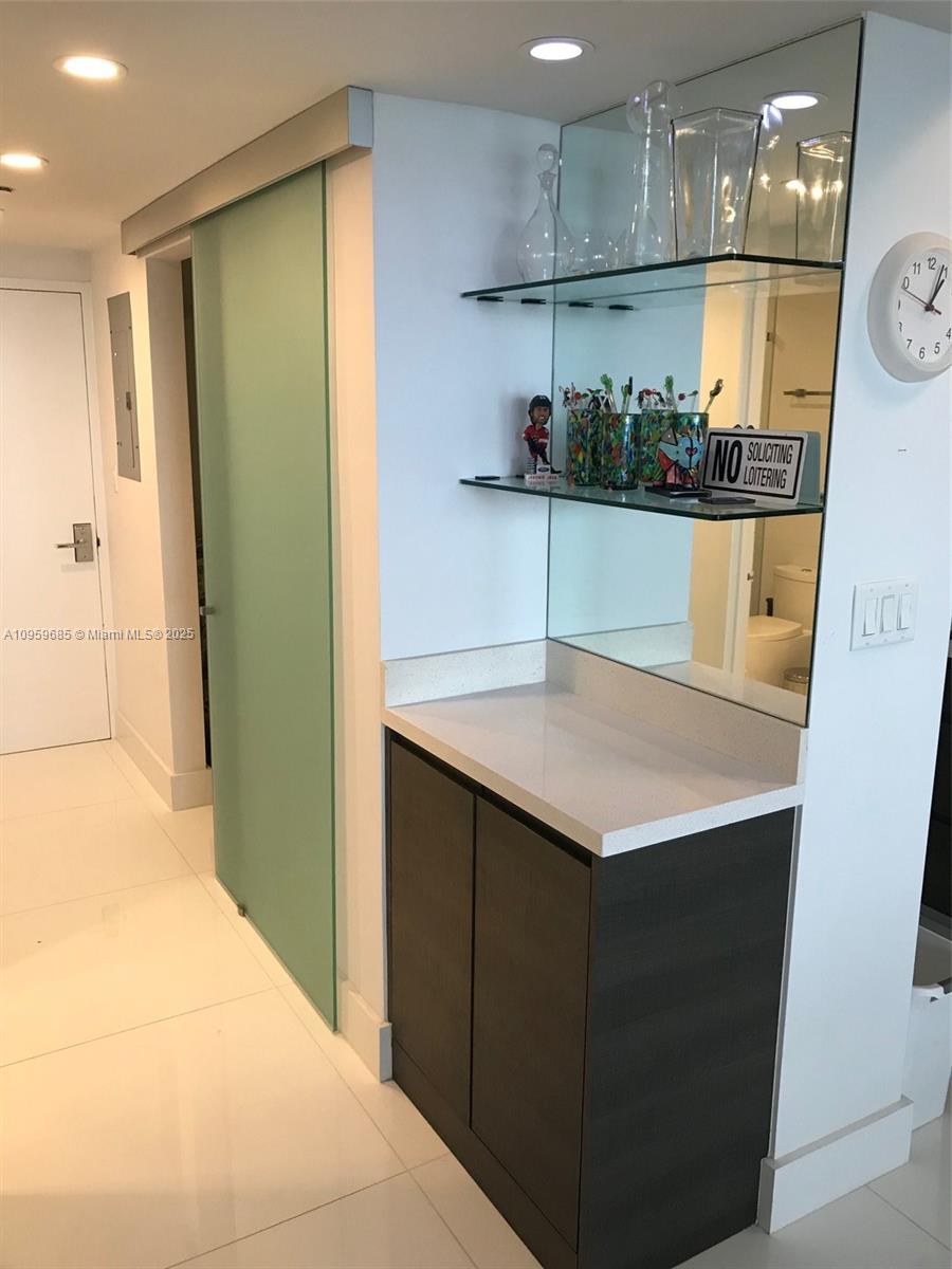 A wet bar with dark cabinets, white countertops, and glass shelving sits beside a sliding door — 100 Lincoln Rd # 719, Miami Beach FL 33139, MIAMI BEACH, FL
