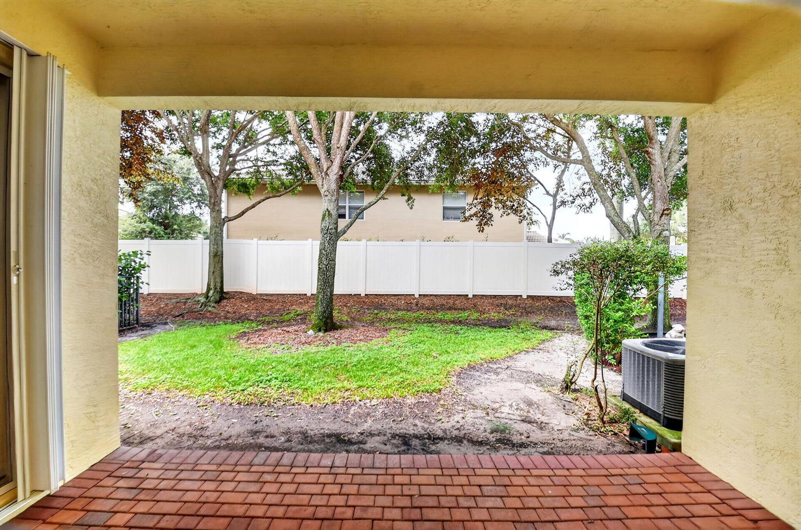 A covered brick patio overlooks a backyard with a lawn, trees, and a white fence — 7662 Spatterdock Drive, Boynton Beach FL 33437, BOYNTON BEACH, FL