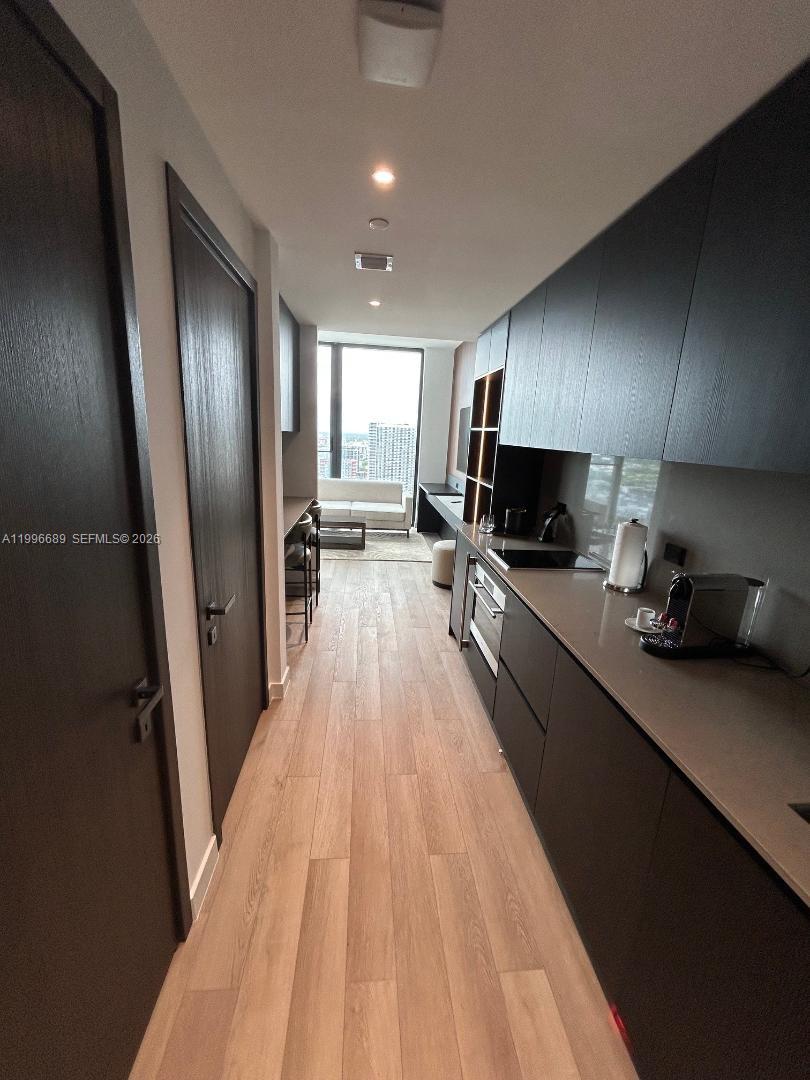 A narrow kitchen with dark cabinetry and light wood flooring leads toward a living area — 20 NE 11 st # 3301, Miami FL 33132, MIAMI, FL