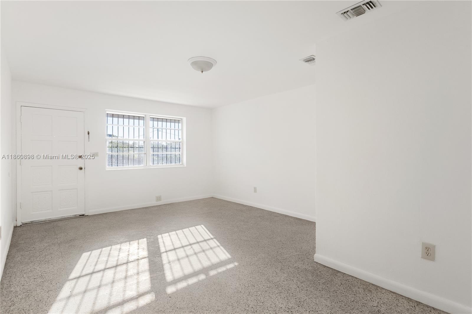 An empty room features white walls, gray carpet, a window with security bars, and a door — 1251 NW 59th St # 3, Miami FL 33142, MIAMI, FL
