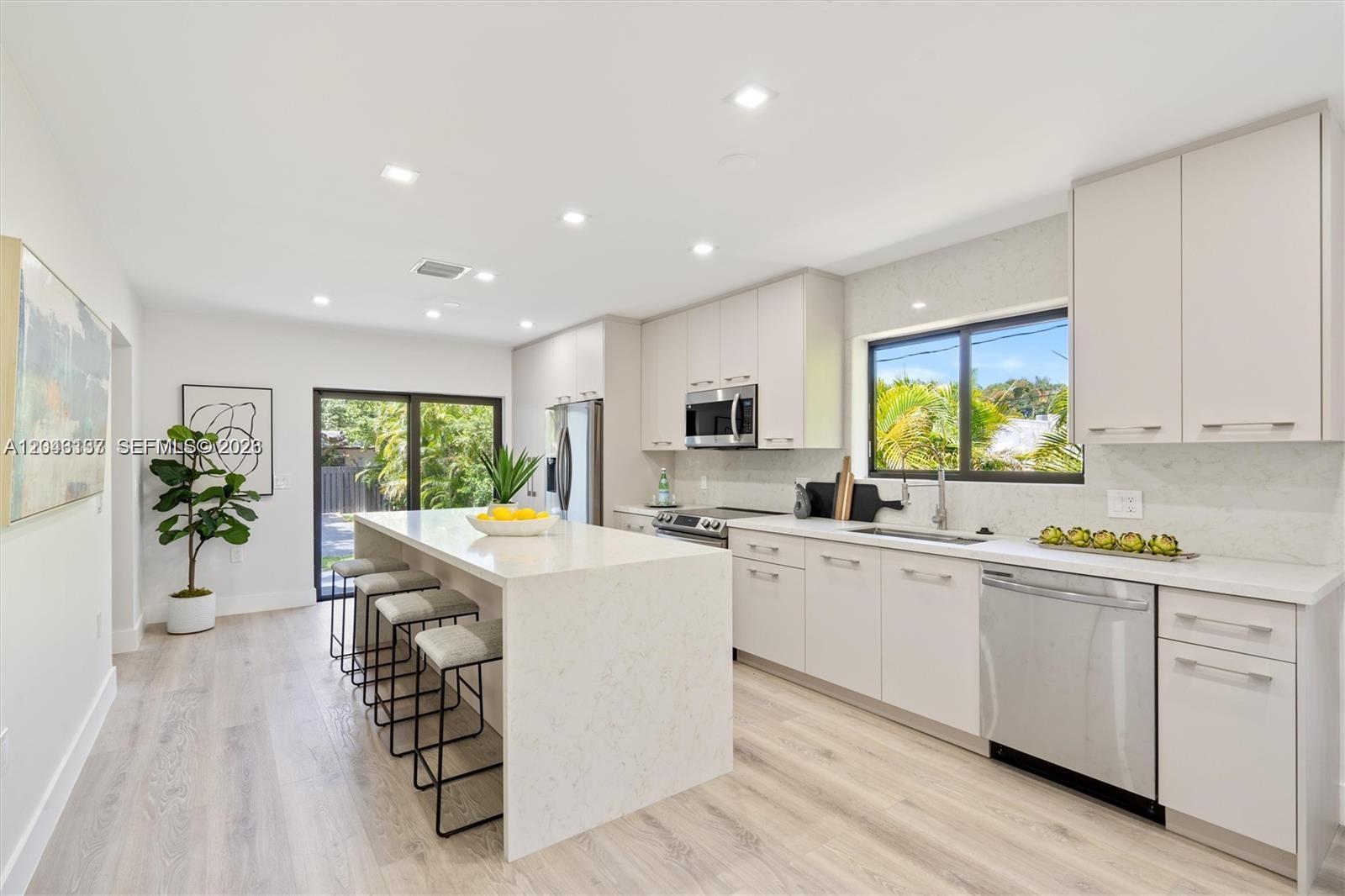 A modern kitchen features white cabinetry, a quartz waterfall island, and light wood-look flooring — 222 SW 3rd Ter, Dania Beach FL 33004, DANIA BEACH, FL