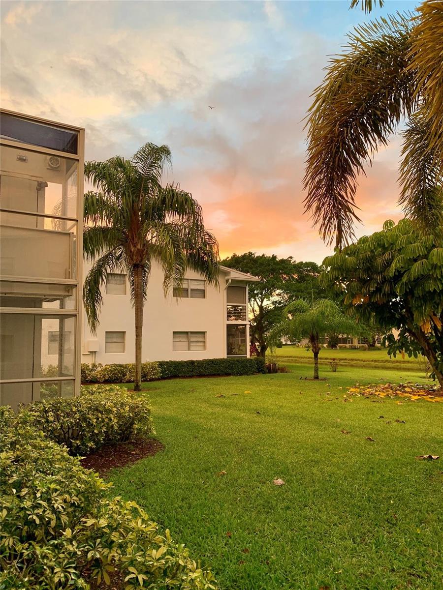 A white multi-story apartment building stands behind a grassy lawn with palm trees at sunset — 236 Prescott-l # 236, Deerfield Beach FL 33442, DEERFIELD BEACH, FL