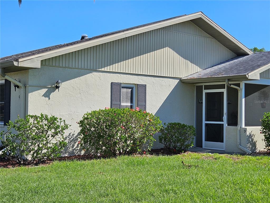 A single-story house exterior features a stucco wall, vertical siding, a screened entryway, and landscaping — 1905 CANTERBURY LANE UNIT E-9, SUN CITY CENTER, FL