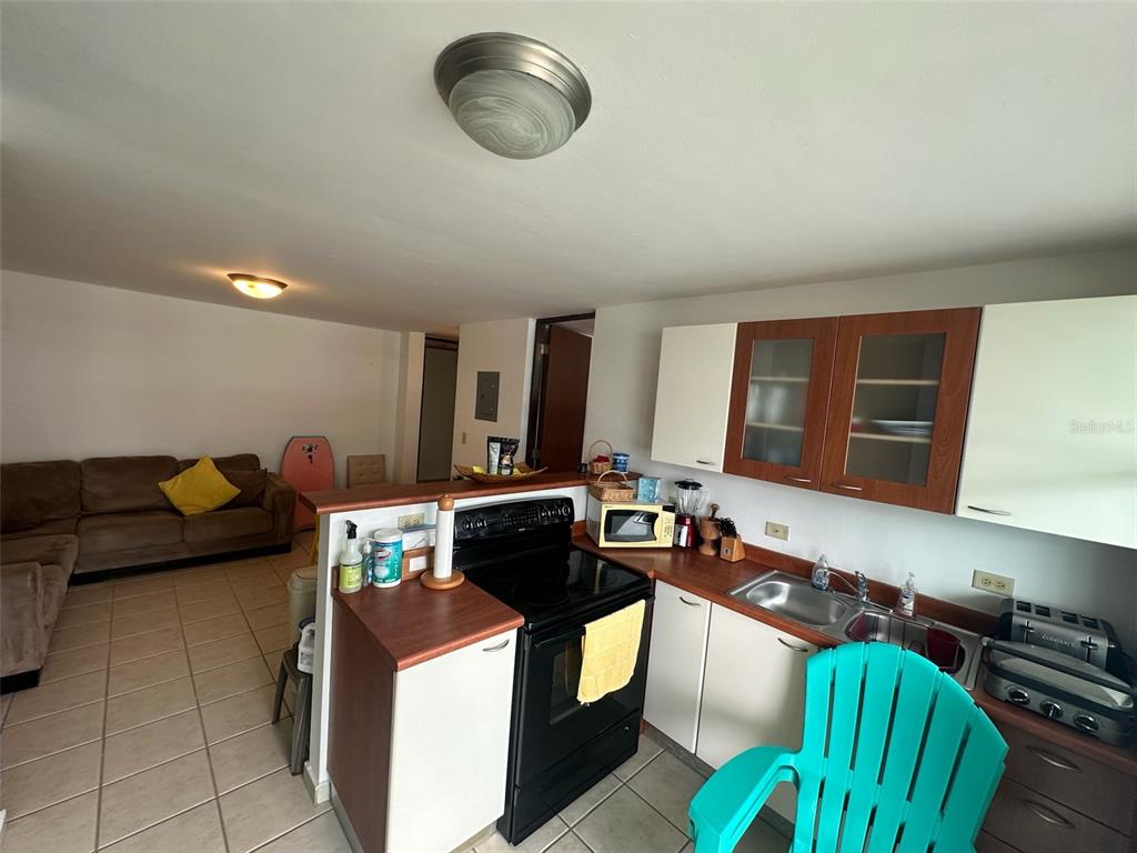 A kitchen with black appliances and wood-toned cabinets opens into a living room with tile floors — Km 7 EL CHARLIE UNIT NE204, LOIZA, PR