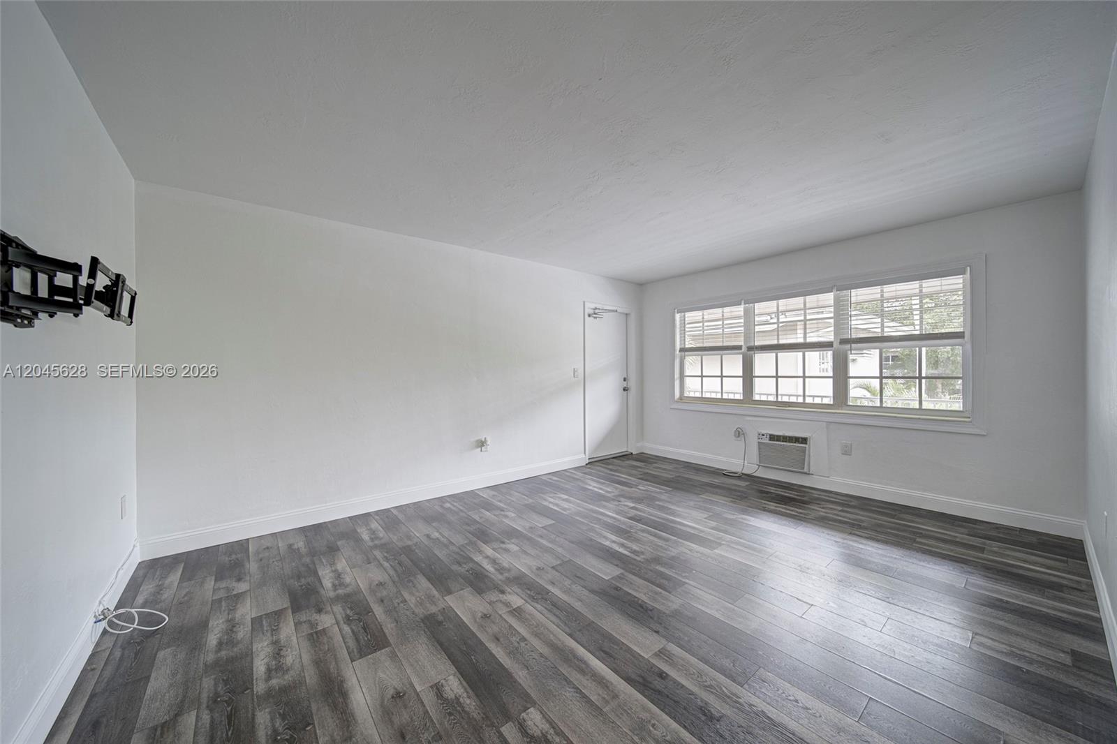An empty room features white walls, gray wood-look flooring, a window, and a wall-mounted TV bracket — 6565 Santona St # B19, Coral Gables FL 33146, CORAL GABLES, FL