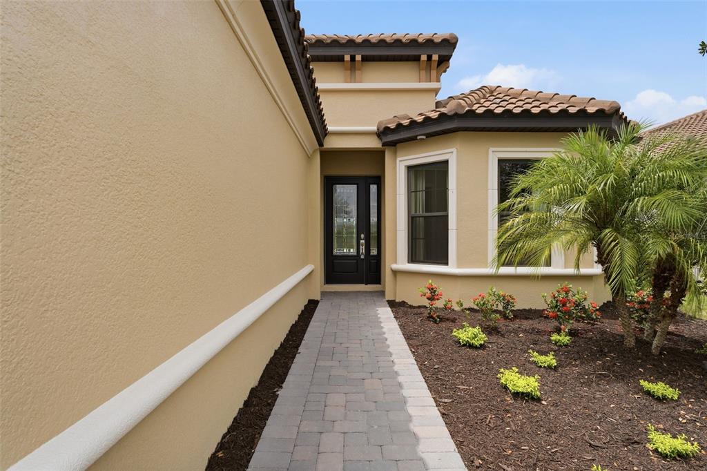 A paved walkway leads to the front entrance of a tan house with tile roofing — 7708 MOONBEAM AVENUE, SARASOTA, FL