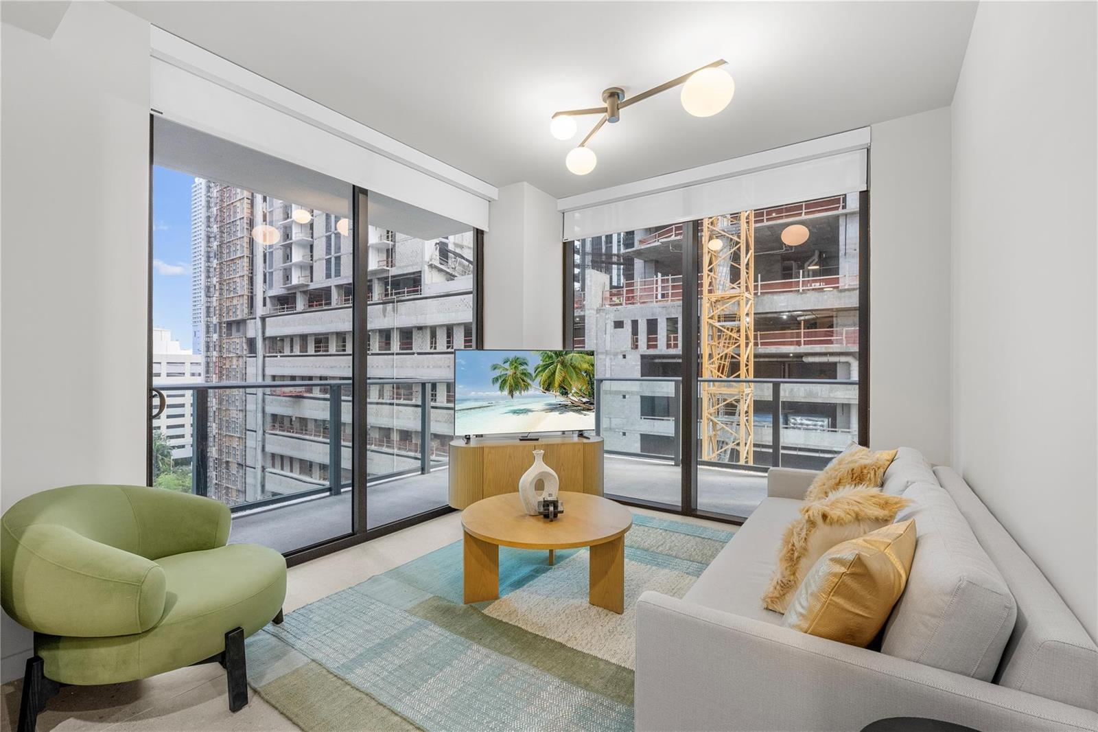 A living room with a sofa, green chair, coffee table, and floor-to-ceiling windows overlooking construction — 11 NE 6th Street # 907, Miami FL 33132, MIAMI, FL