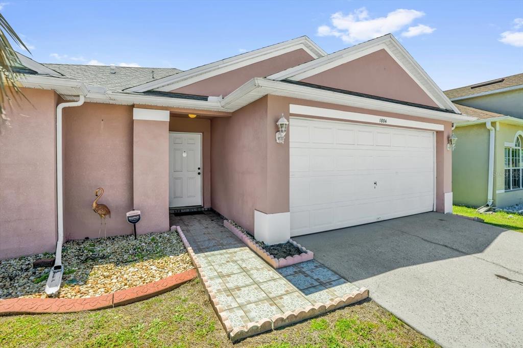 A pink single-story house features a white garage door and a tiled walkway to the entrance — 1804 BAGUETTE COURT, KISSIMMEE, FL