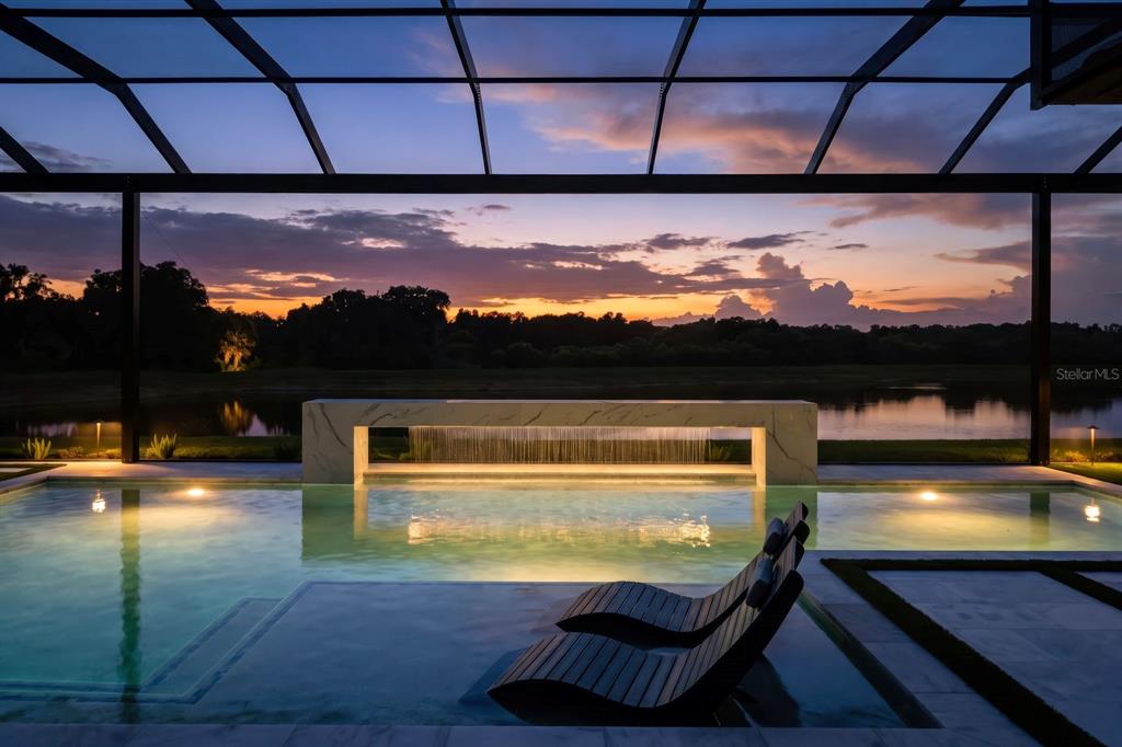 A swimming pool with a waterfall feature and lounge chairs overlooks a lake at sunset — 2428 WATERFRONT CIRCLE, SARASOTA, FL
