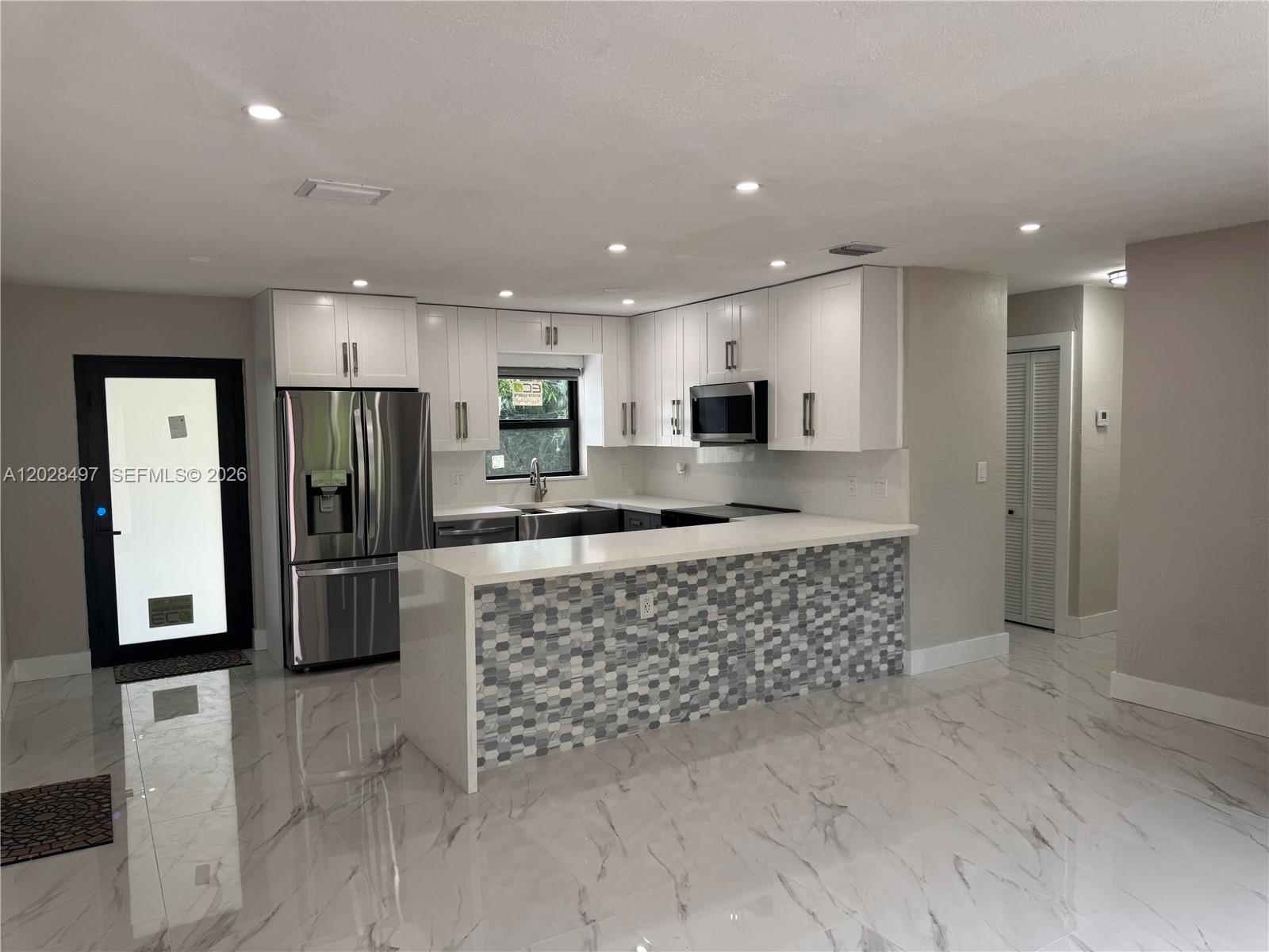 A modern kitchen features white cabinets, stainless steel appliances, a tiled peninsula, and marble-look flooring — 919 NE 4th Ct, Hallandale Beach FL 33009, HALLANDALE BEACH, FL