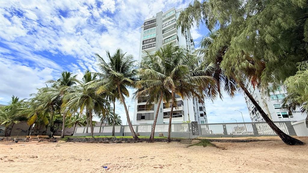 A tall white apartment building stands behind a sandy beach with several palm trees — 37 ISLA VERDE AVE. UNIT 203, CAROLINA, PR