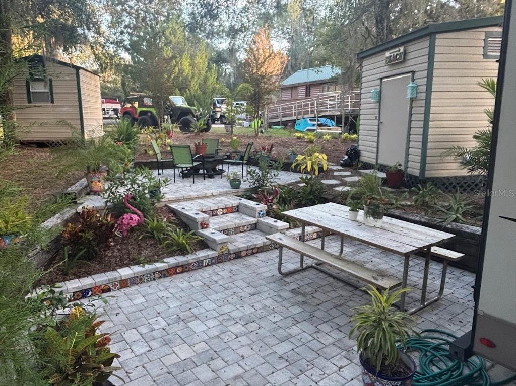 A backyard patio features paver flooring, a picnic table, tiered steps, and two storage sheds — 25244 NE 143RD  ST UNIT 180, SALT SPRINGS, FL