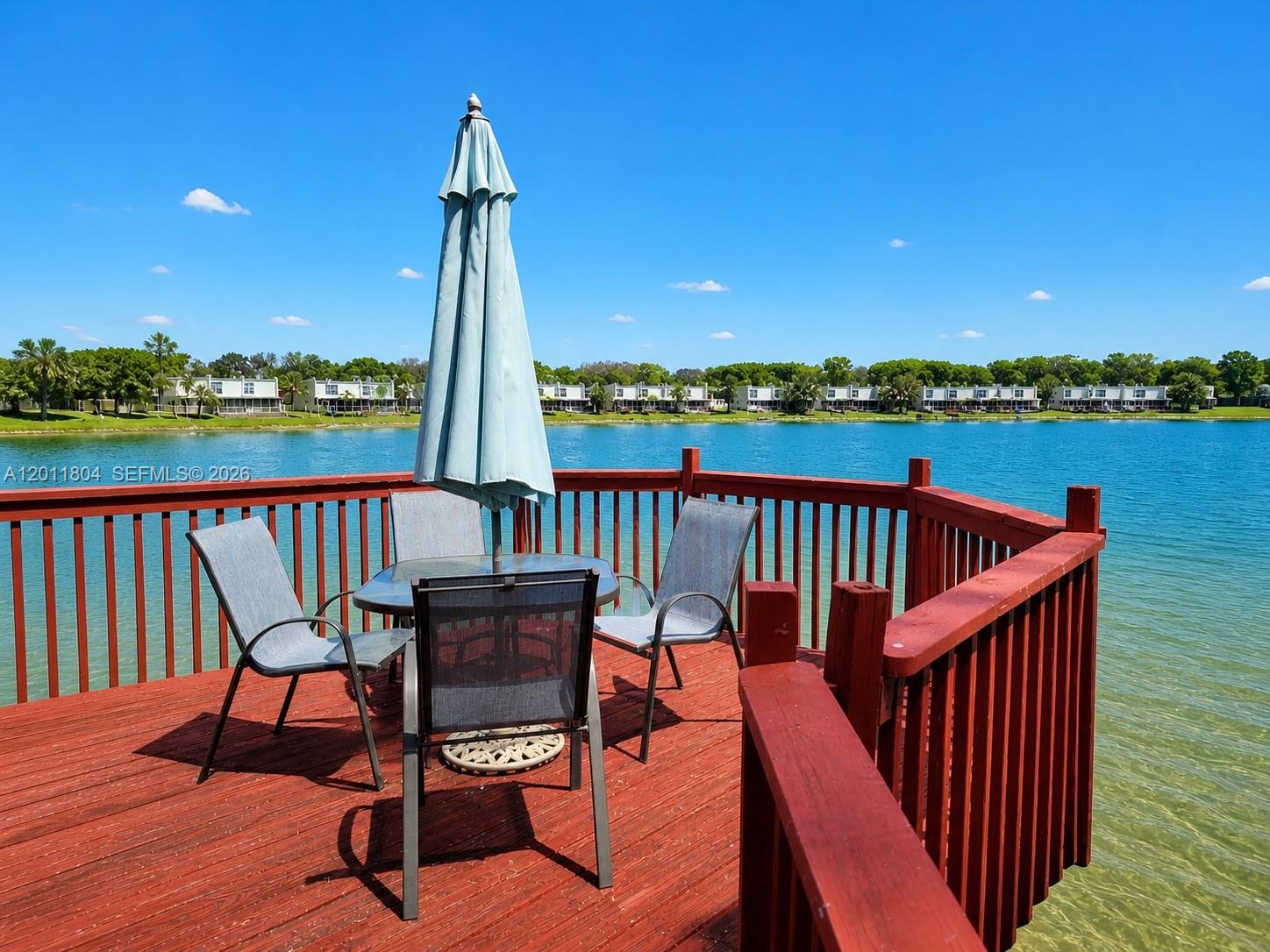 A red wooden deck with patio furniture overlooks a lake under a clear blue sky — 14020 SW 83rd St, Miami FL 33183, MIAMI, FL