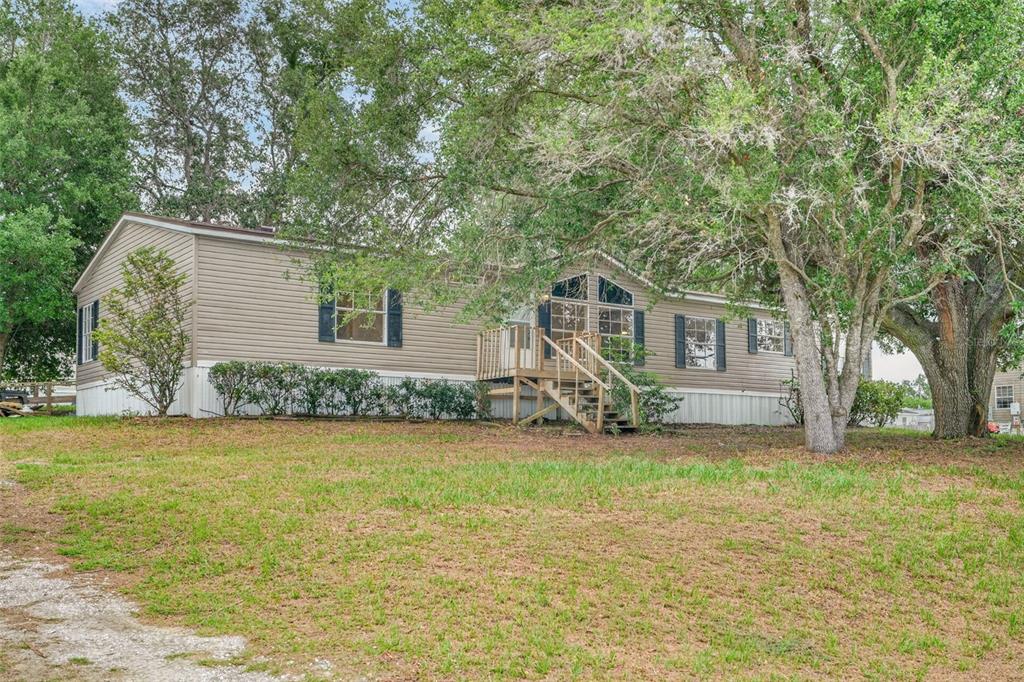 A tan manufactured home with a wooden front deck sits on a grassy, tree-filled lot — 14199 SE 59TH COURT, SUMMERFIELD, FL