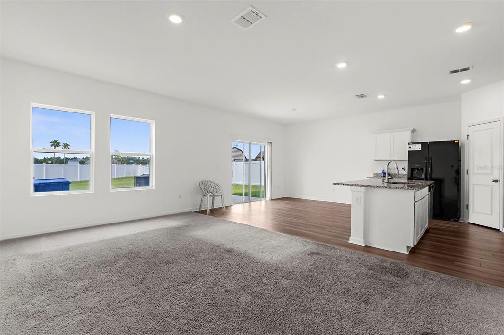 An open-concept living room features gray carpeting, white walls, and a kitchen with dark appliances — 1038 KELLY FERN LOOP, RUSKIN, FL