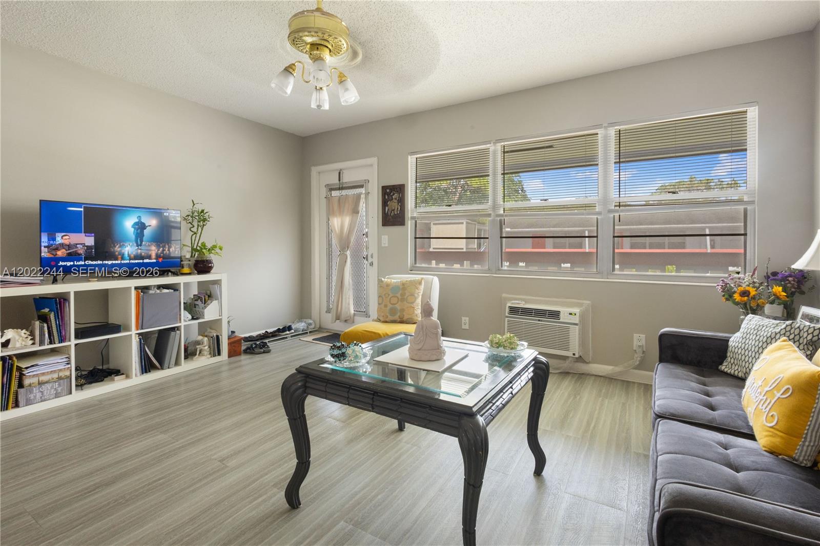 A living room features light wood-look flooring, a glass-top coffee table, and a television console — 75 Ventnor D # 75, Deerfield Beach FL 33442, DEERFIELD BEACH, FL