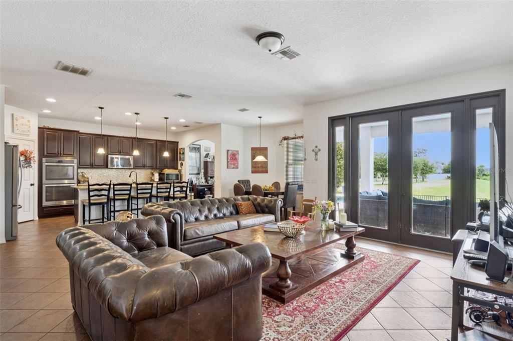 An open-concept living room and kitchen featuring dark leather sofas, tile floors, and glass doors — 9189 BUSACO PARK WAY, WINTER GARDEN, FL
