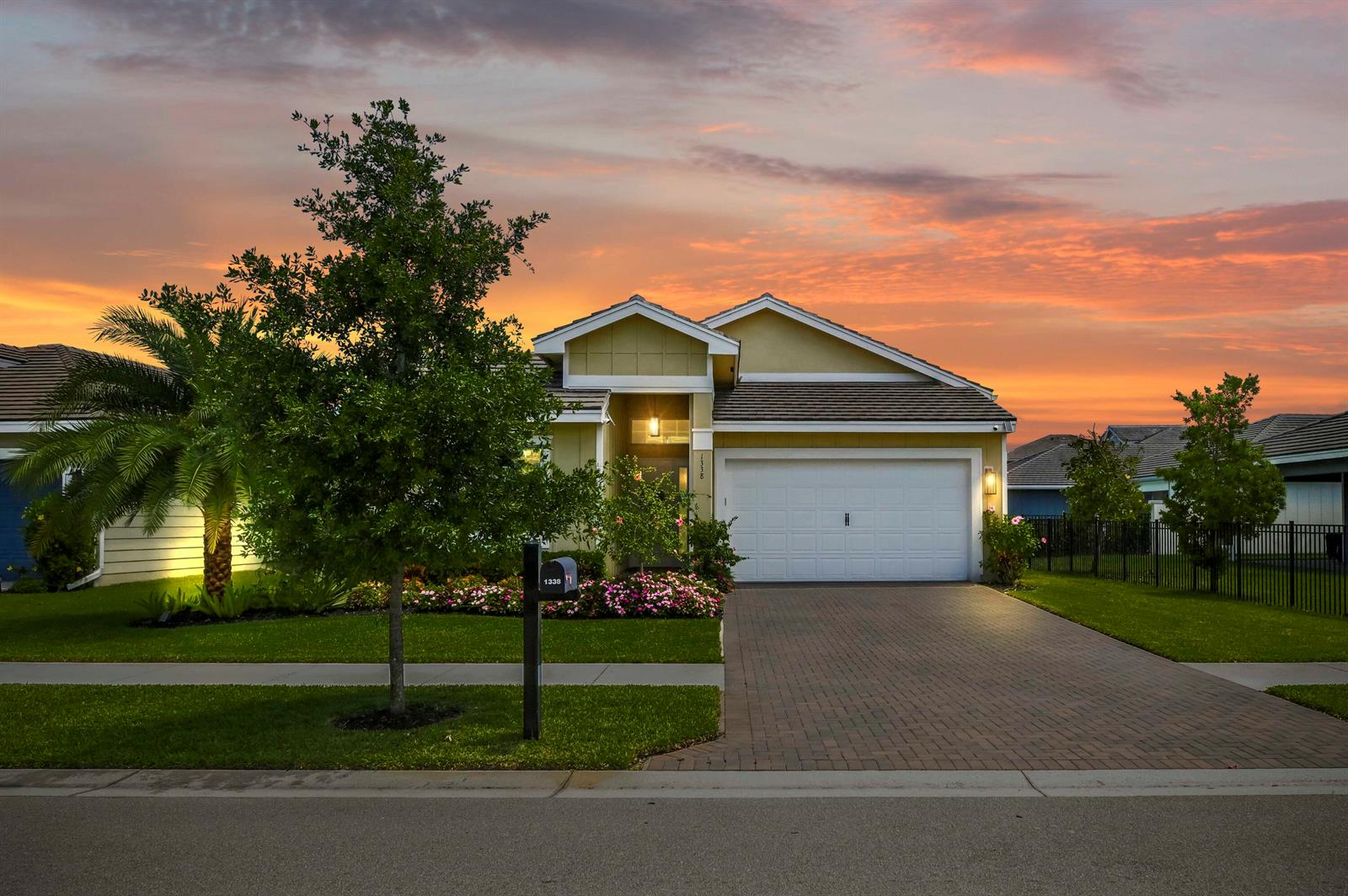 A single-story house with a white garage, paved driveway, and a palm tree at sunset — 1338 Harvester Crossing, Loxahatchee FL 33470, LOXAHATCHEE, FL