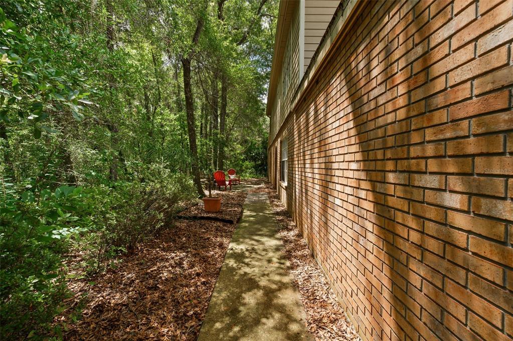 A concrete walkway runs alongside a brick house exterior next to a wooded area — 5261 SW 97TH DRIVE, GAINESVILLE, FL