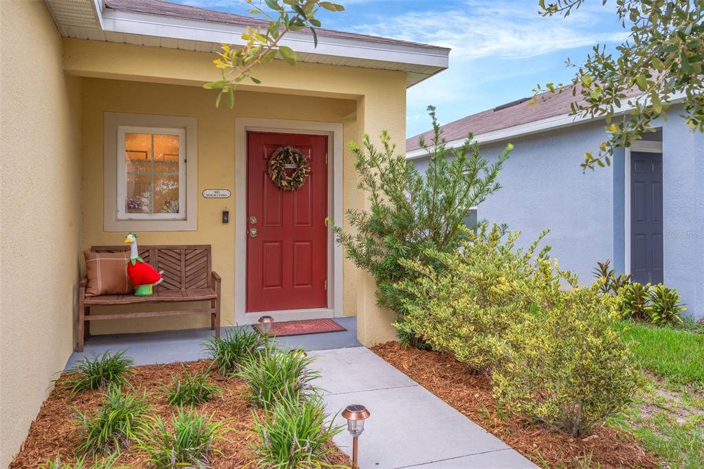 A red front door with a wreath sits under a covered porch next to a bench — 35088 JOMAR AVENUE, ZEPHYRHILLS, FL
