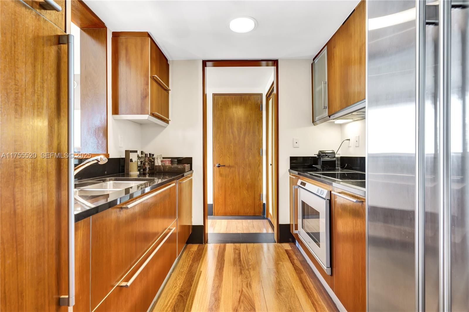 A galley kitchen features wood cabinetry, black countertops, stainless steel appliances, and hardwood flooring — 101 20th St # 1803, Miami Beach FL 33139, MIAMI BEACH, FL