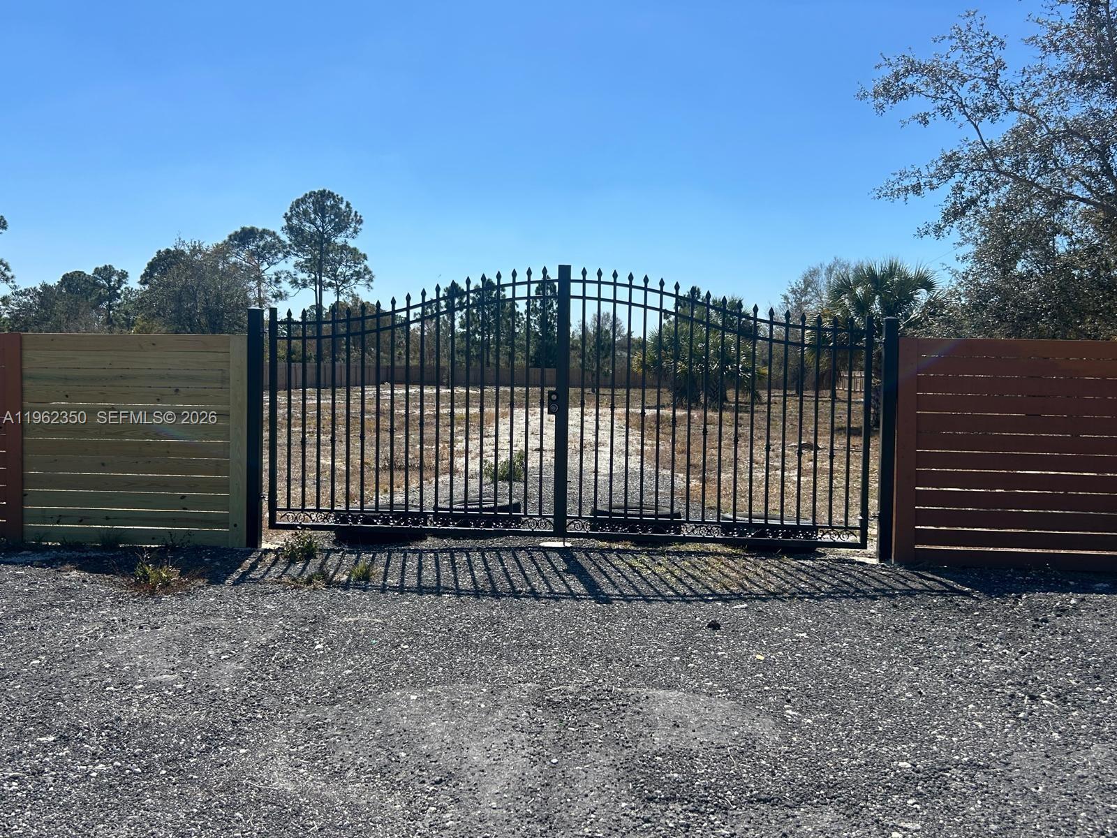 A black metal gate stands between two wooden fences on a gravel lot — 648 Hunting Club Ave, Clewiston FL 33440, CLEWISTON, FL