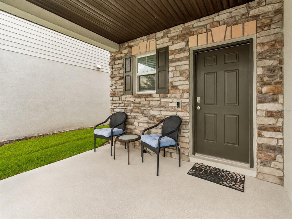 A covered concrete patio features stone veneer walls, a dark door, and two outdoor chairs — 7840 ABBY BROOKS CIRCLE, WESLEY CHAPEL, FL