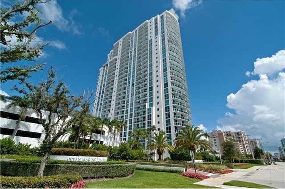 A tall, modern residential high-rise building stands behind a landscaped lawn and parking structure — 1945 S Ocean Drive # 910, Hallandale Beach FL 33009, HALLANDALE BEACH, FL