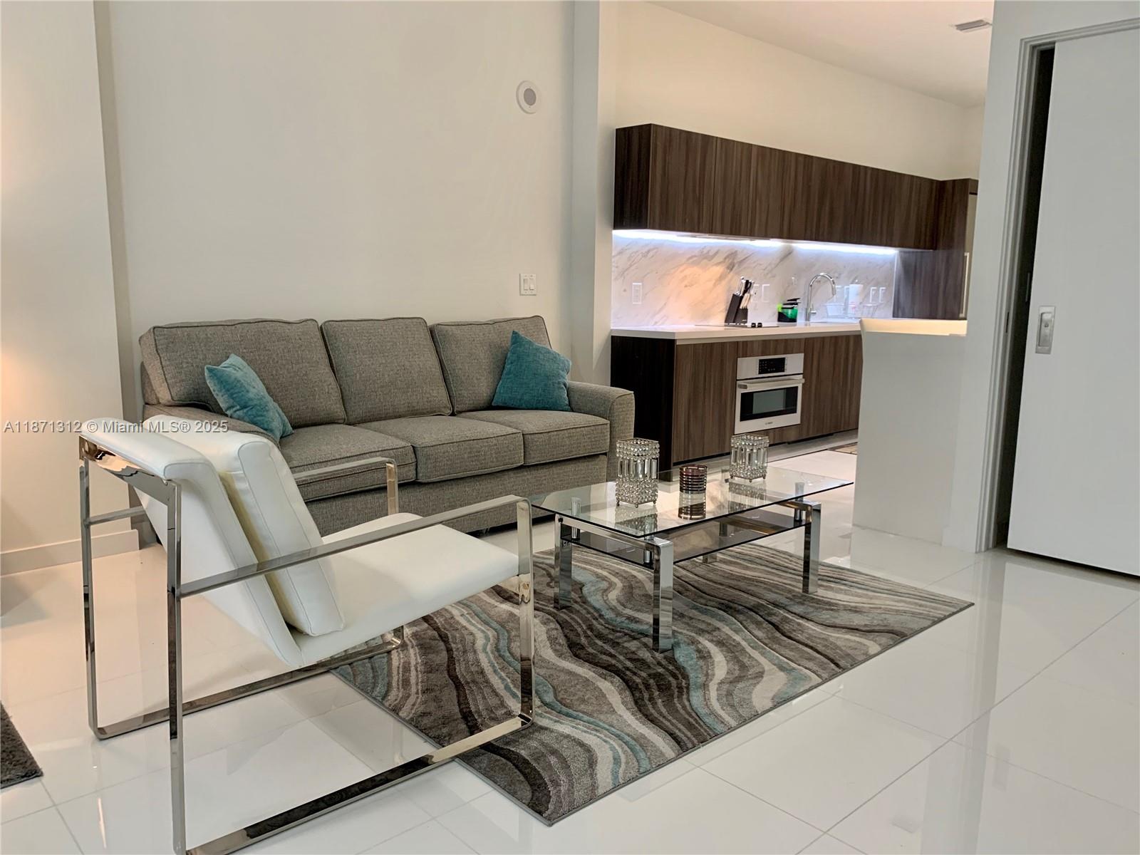 A modern living room features a grey sofa, white accent chair, and an open kitchen — 851 NE 1 Ave # 502, Miami FL 33132, MIAMI, FL