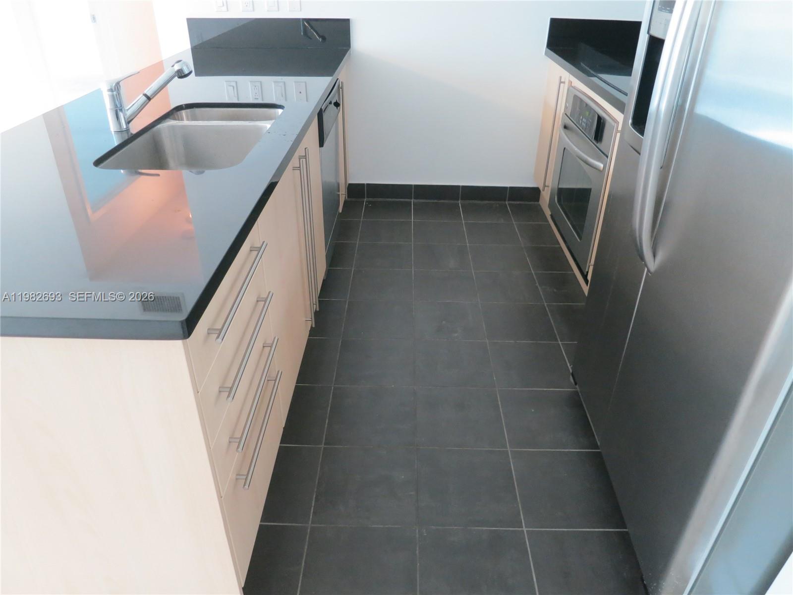 A galley kitchen features light wood cabinets, black countertops, stainless steel appliances, and dark tile — 1111 SW 1 AV, Miami FL 33130, MIAMI, FL