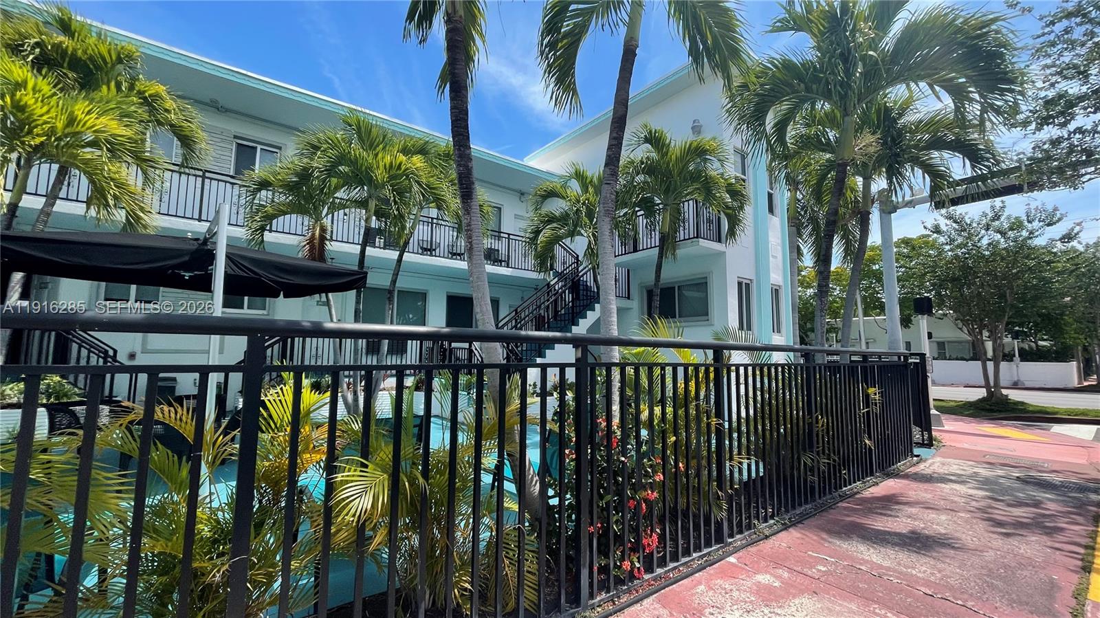 A two-story light blue apartment building features a pool, palm trees, and black metal fencing — 8100 Harding Ave # 508, Miami Beach FL 33141, MIAMI BEACH, FL