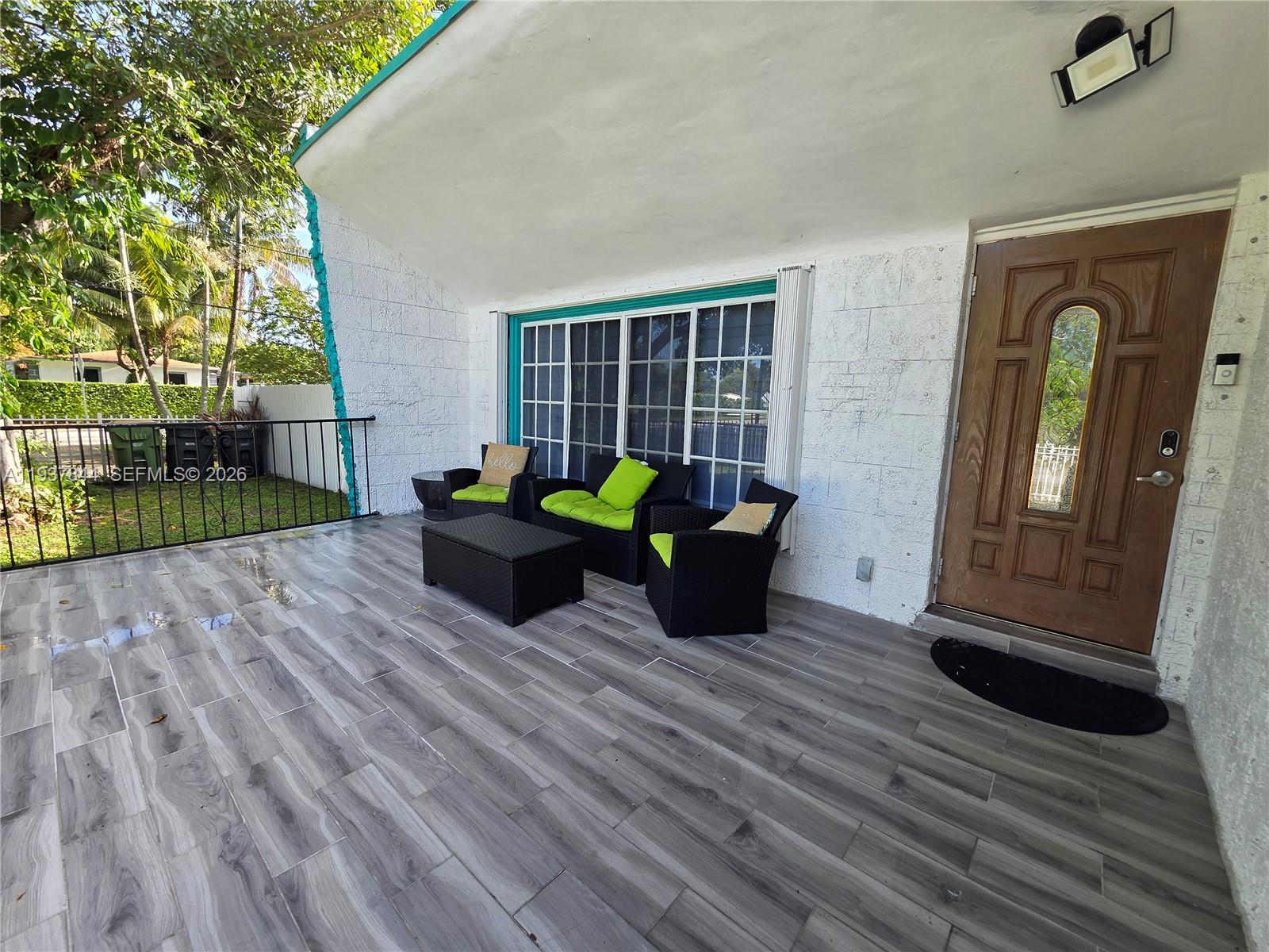 A covered patio features gray wood-look tile flooring, outdoor furniture, and a wooden front door — 13895 NE 11th Ave, North Miami FL 33161, NORTH MIAMI, FL