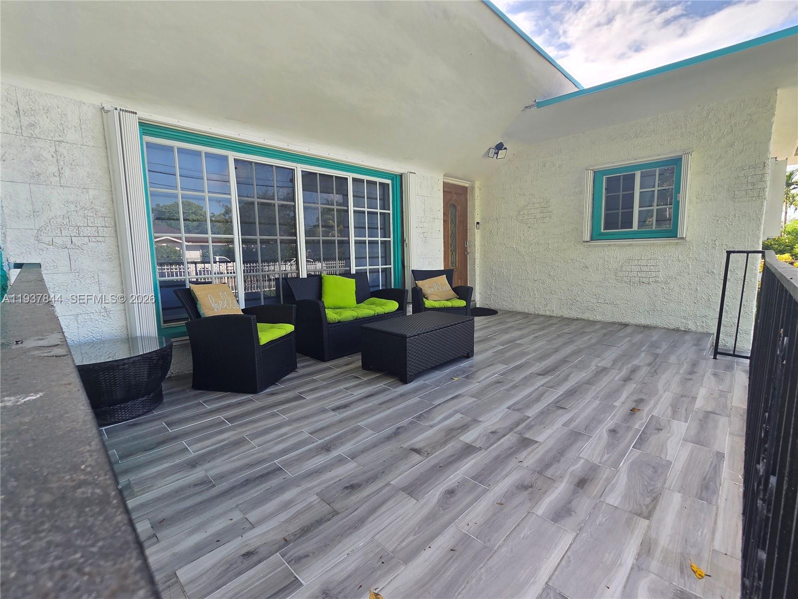 A covered patio features gray wood-look tile flooring, white stucco walls, and black wicker furniture — 13895 NE 11th Ave, North Miami FL 33161, NORTH MIAMI, FL