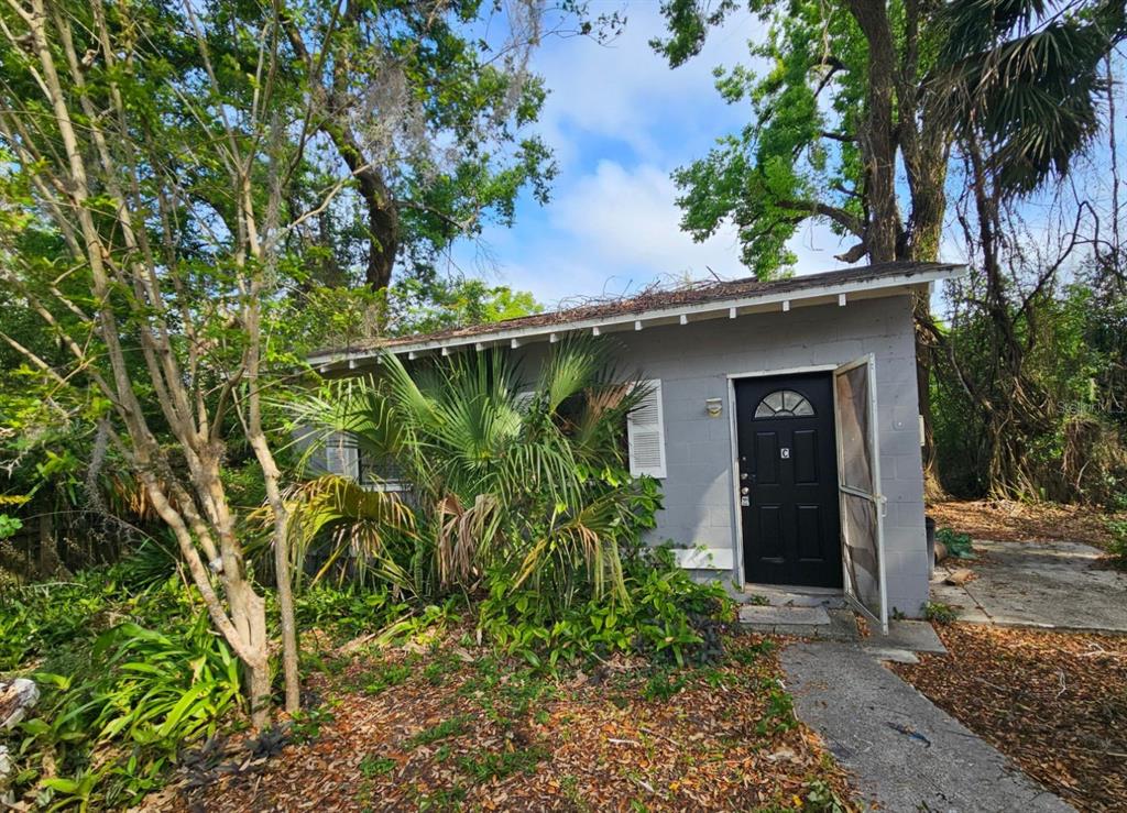 A small, gray concrete block building with a black front door and surrounding overgrown vegetation — 1504 E 99TH AVENUE UNIT C, TAMPA, FL