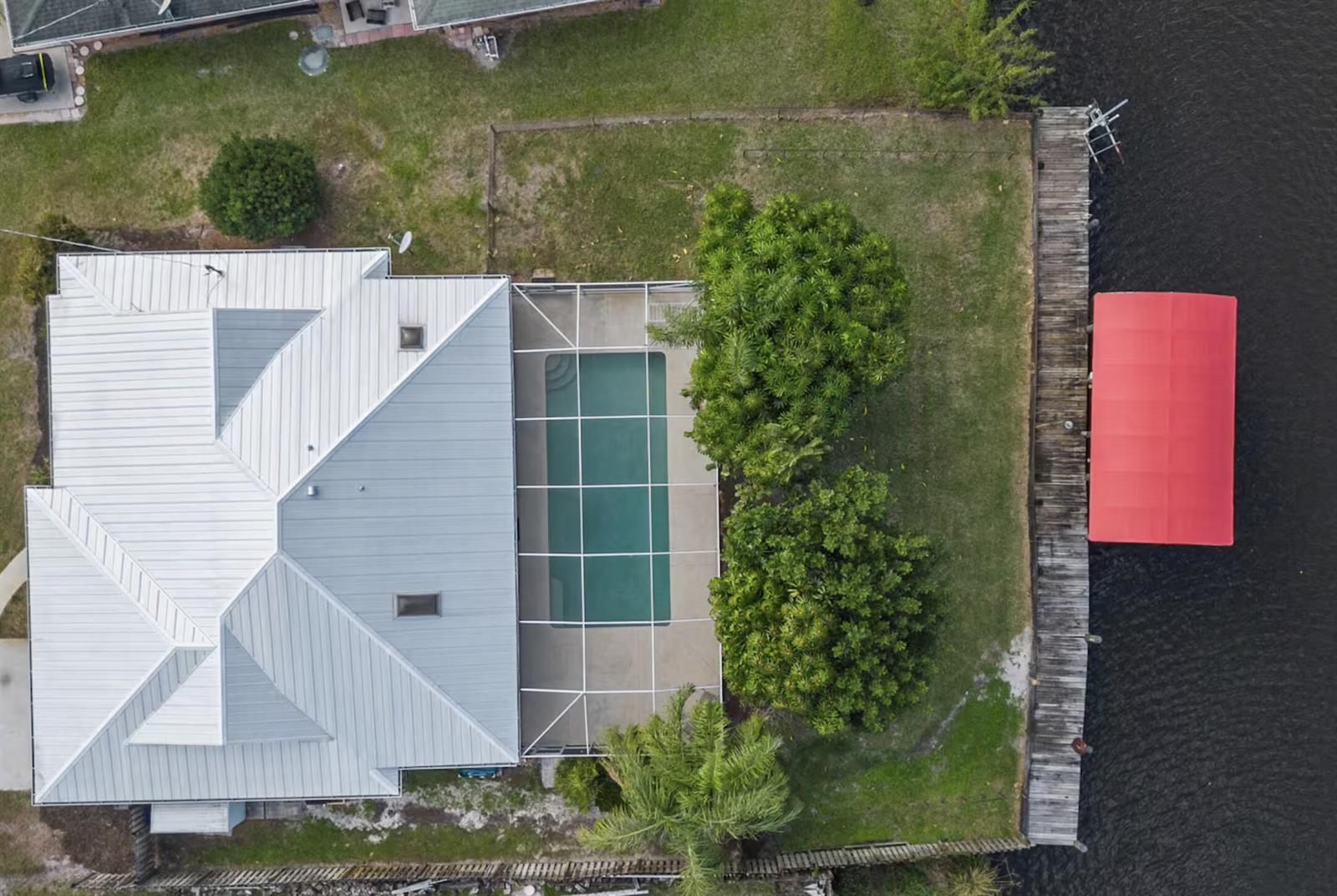 An aerial view shows a house with a metal roof, pool enclosure, and wooden dock — PORT ST. LUCIE, FL
