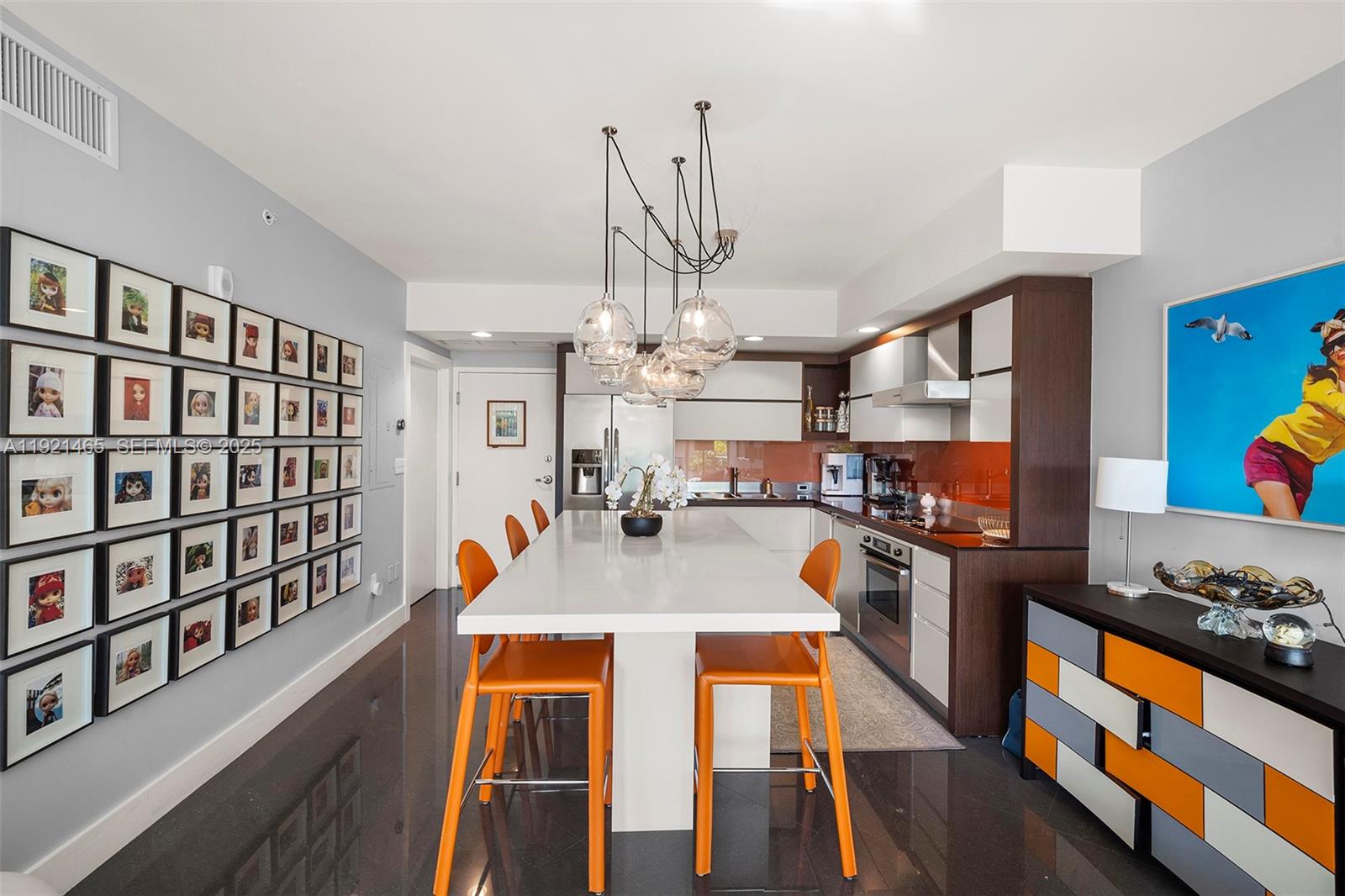 A modern kitchen features a white island with orange stools and a wall of framed photos — 2155 Washington Ct # 205, Miami Beach FL 33139, MIAMI BEACH, FL