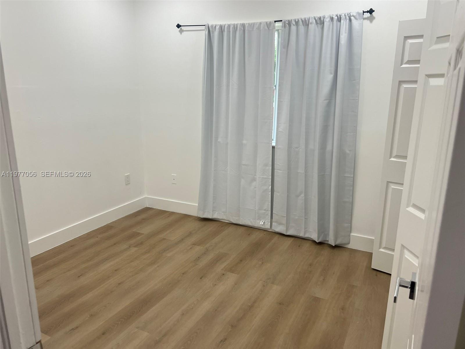 An empty bedroom features light wood-look flooring, white walls, and gray curtains over a window — 2511 NE 41st Ter, Homestead FL 33033, HOMESTEAD, FL