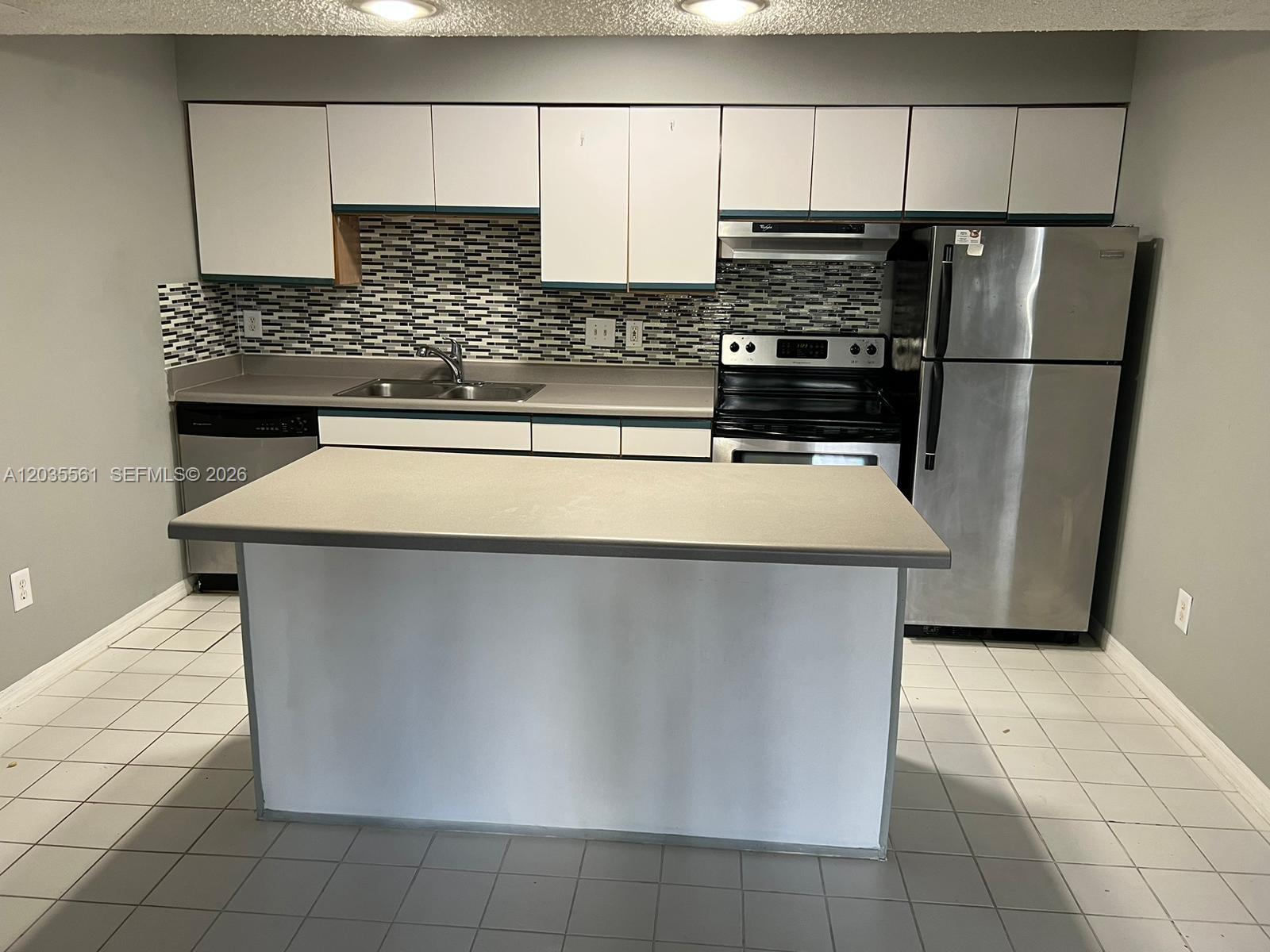 A kitchen features white cabinets, a gray island, stainless steel appliances, and a tiled backsplash — 3985 W Mcnab Rd # A103, Pompano Beach FL 33069, POMPANO BEACH, FL