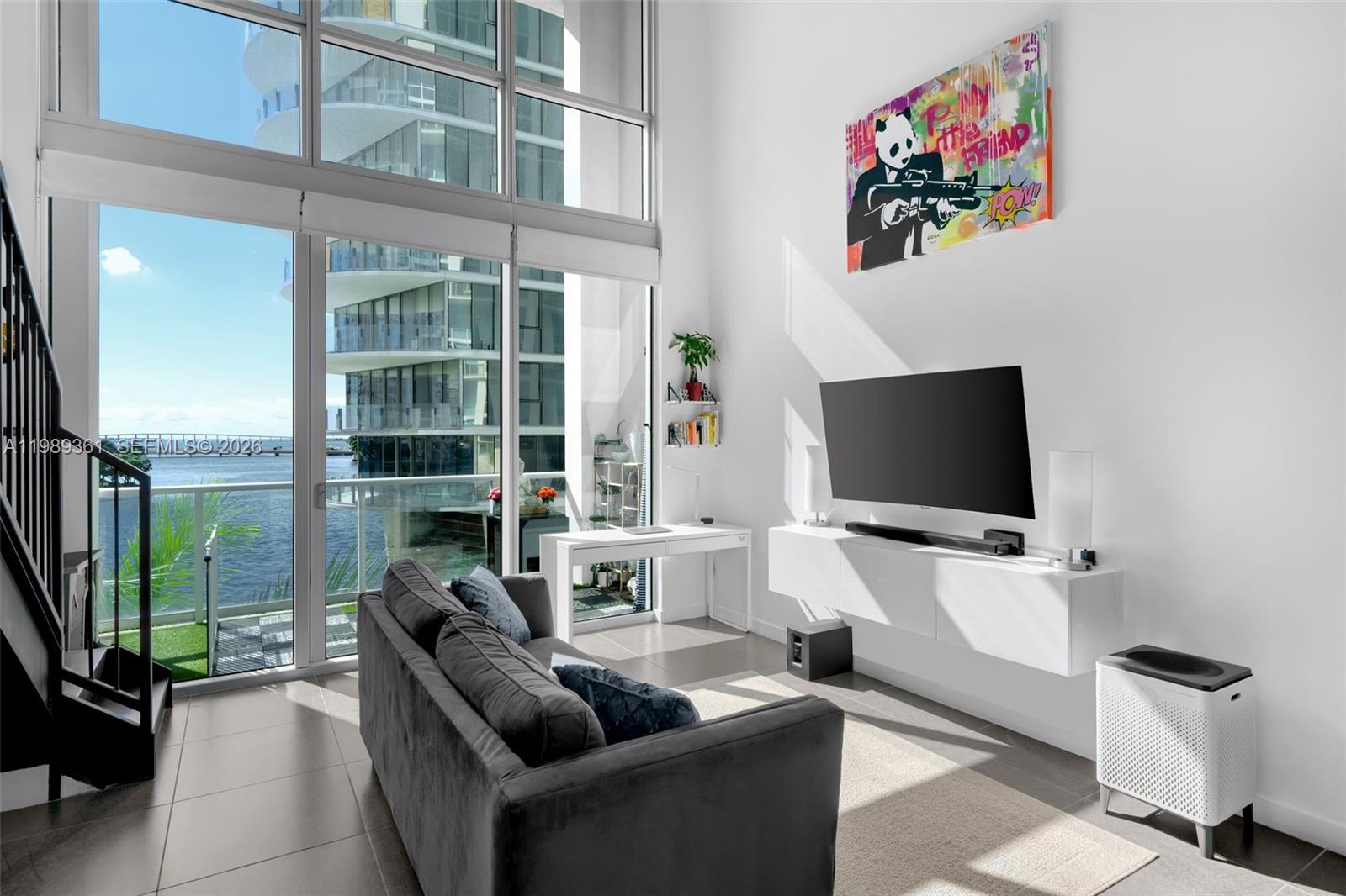 A modern living room features a grey sofa, wall-mounted television, desk, and floor-to-ceiling windows — 300 S BISCAYNE BLVD # 410, Miami FL 33131, MIAMI, FL