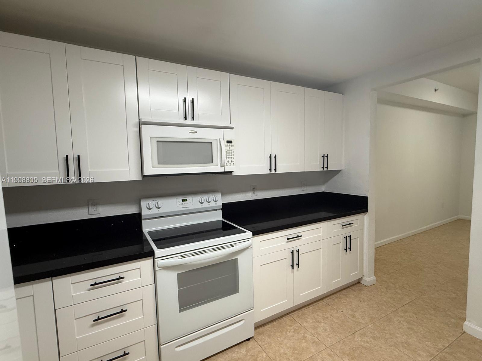 A kitchen features white shaker cabinets, black countertops, and white appliances on a tiled floor — 11603 NW 89th St # 215, Doral FL 33178, DORAL, FL