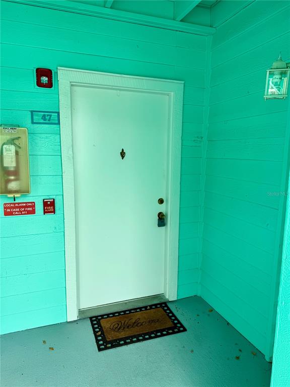 A white front door with a welcome mat is set against bright teal exterior walls — 455 ALT 19 S UNIT 47, PALM HARBOR, FL