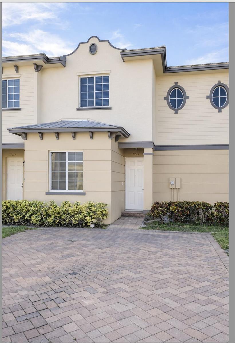 A two-story townhouse exterior with a paved driveway, tan stucco walls, and two white doors — 1219 Lucaya Drive, Riviera Beach FL 33404, RIVIERA BEACH, FL