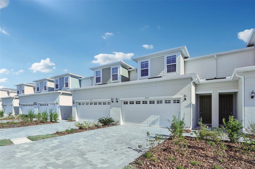 A row of modern townhomes with light-colored stucco, attached garages, and paved driveways under blue skies — 24101 MANUMA STREET UNIT Seminole 2491, ST CLOUD, FL