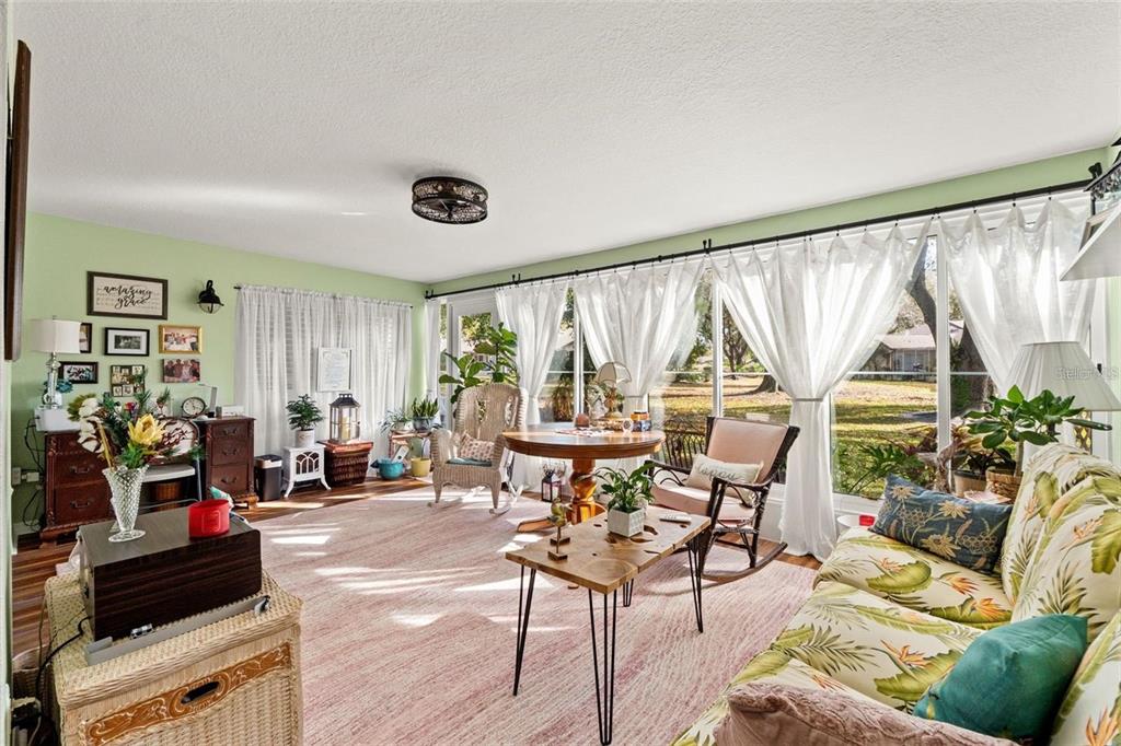 A sunroom features a floral sofa, a wooden coffee table, and large windows with white curtains — 7427 HEATHER WALK DRIVE, WEEKI WACHEE, FL