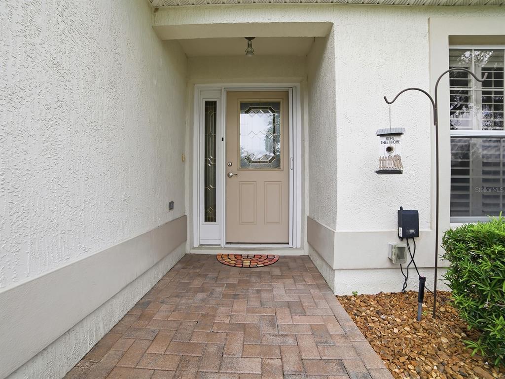 A paved walkway leads to a beige front door with a sidelight and storm door — 7290 SE 172ND LEGACY LANE, THE VILLAGES, FL