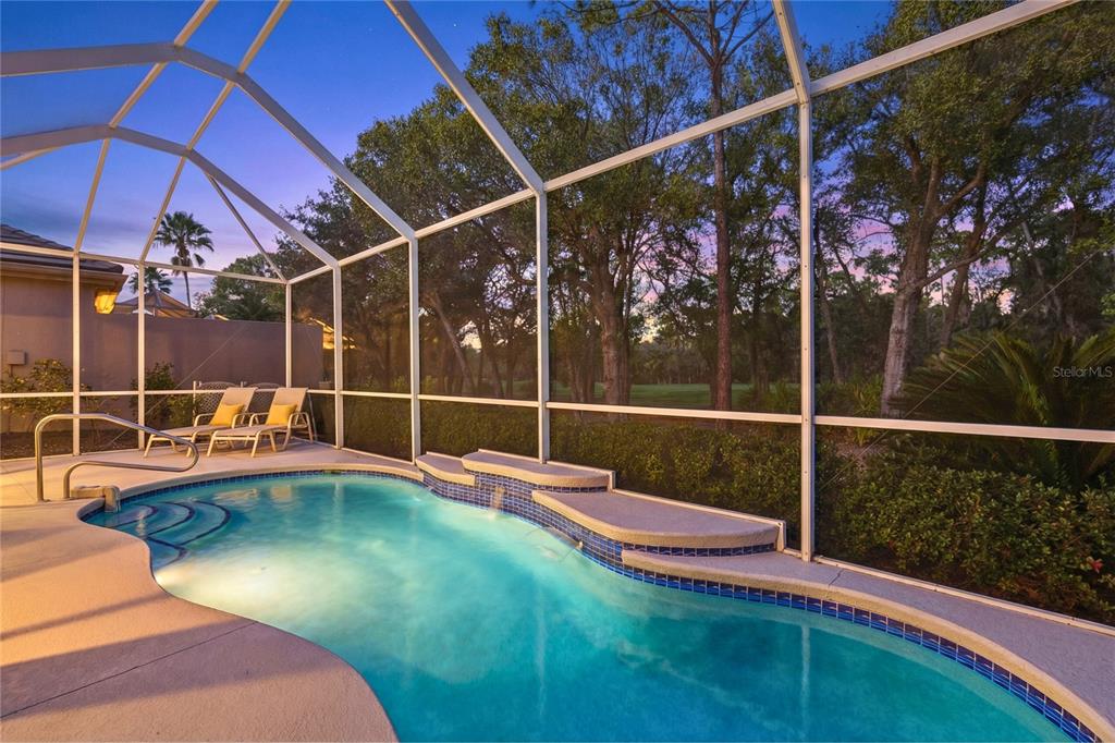 A swimming pool with a water feature is enclosed by a screened lanai at dusk — 8733 49TH TERRACE EAST, BRADENTON, FL